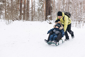 Fototapeta na wymiar Father with backpack and little sons walking together in winter snowy forest. Happy man and joyful boys sledding and having fun together. Wintertime activity outdoors. Concept of local travel
