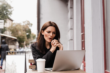 Shocked girl is winning bet in online casino and sitting with laptop in cafe outdoor. Cheerful surprised woman smiling and looking at amount of income earnings in online casino and rejoicing outside.