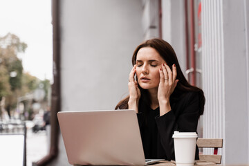Woman manager with strong headache working on laptop online remotely outside of cafe. Sad girl with laptop and cup of coffee got migraine touches her head because of pain.