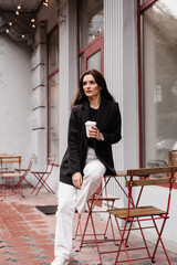 Confident young business woman with cup of coffee is posing outside of cafe on terrace. Cheerful girl is weared casual business style is posing near cafe.