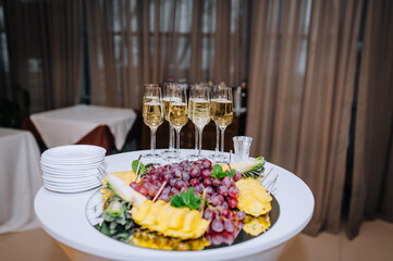 Glass wine glasses with champagne, sparkling drink stand on a round wooden white table with fruits, dessert and dishes in a restaurant. Photography, banquet, wedding.
