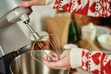 Woman in festive christmas sweater preparing dough for cookies at home kitchen. Female hands use electric mixer. Modern kitchen household appliances
