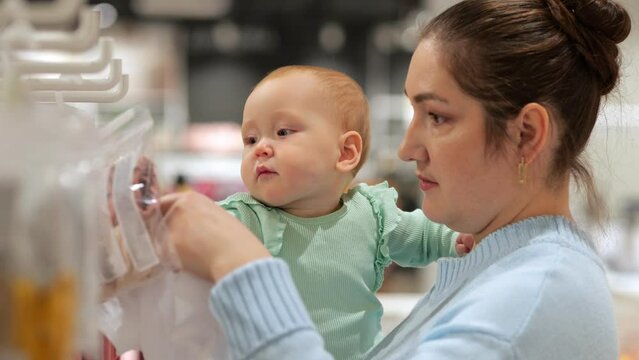 Baby girl with serious expression helps mother choose outfit for home. Child with amused and puzzled expression enjoys shopping with parent closeup