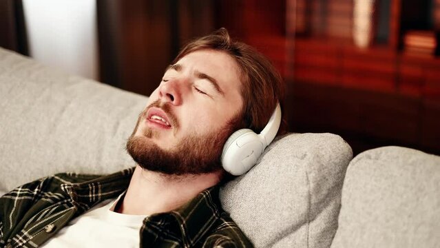 Portrait of attactive young barded man with closing eyes listening to music while laying on sofa in the living room with warm sunset light. Smiling male enjoying leisure time at home alone.