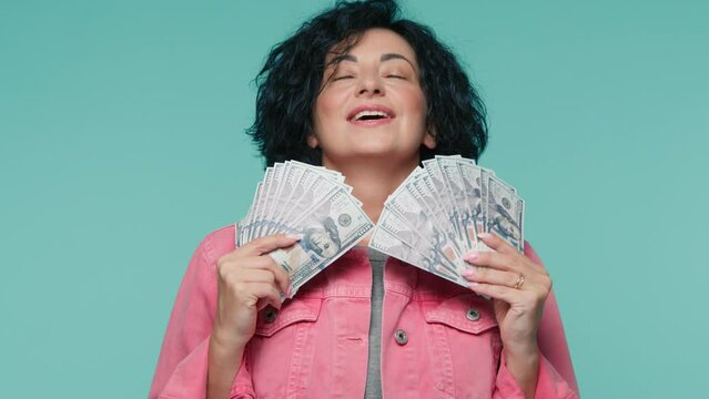 Rich elegant woman 40s in pink jacket smells the fan of dollars banknotes hold in both hands. Spending money on useless thing, isolated blue background. Studio shot happy woman with lots of money