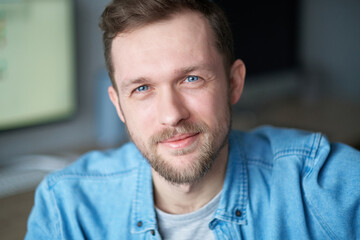 Handsome man in denim shirt, positive emotion with computer monitor on background. Portrait of cheerful bearded male software engineer sitting at working place in office looking at camera