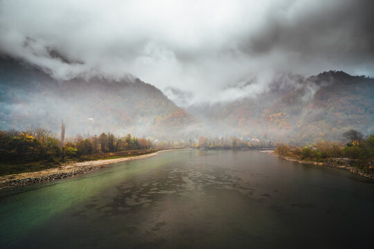 Lake or river with mountains in low clouds and fog. Beautiful autumn travel landscape with hills in mist.