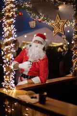 
Portrait von einem verkleideten Weihnachtsmann, der auf einem Weihnachtsmarkt von Lichtern umgeben...
