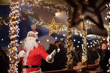 Portrait von einem verkleideten Weihnachtsmann, der auf einem Weihnachtsmarkt von Lichtern umgeben...