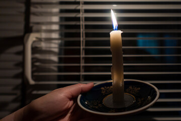 Burning candle in the hands against the background of a window with blinds (close-up). Power outage...