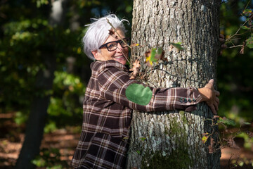 older happy nature mature gray haired woman with glasses, in fifties in sixties hugging a tree...