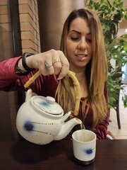young caucasian geisha handmaid pours herself chai tea with teapot in front of bamboo