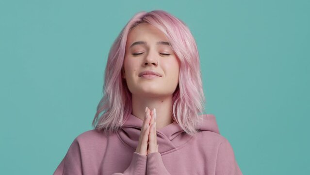 Pleading funny young student woman 20s years old with pastel pink hair color wears pink hoodie hold hands folded in prayer begging about something making wish isolated on blue background slow motion