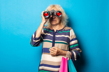 Old woman holding shopping bags and looking through binoculars on blue background