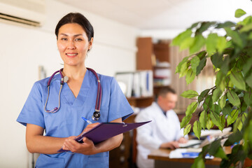 Positive asian doctor woman wearing uniform standing in medical office with clipboard