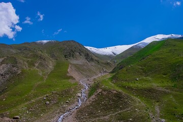 Mountain pass road between two green hills with the rill continuing until covered by snow mountains