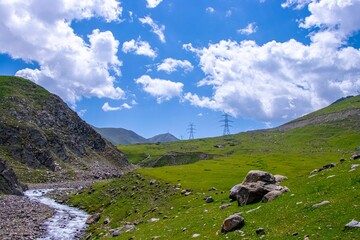 Electric towers stand on the green hillside which crossing by mountain rill surrounded by rocky hill
