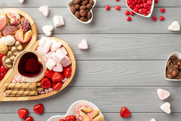 Valentine's Day concept. Top view photo of wooden heart shaped serving tray glass of beverage saucers with sweets candies chocolate cookies on grey wooden desk background with copyspace in the middle
