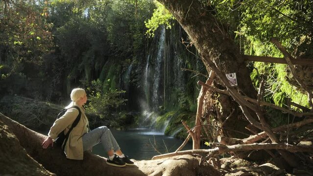 A cinematic shot of a beautiful female tourist sitting on a tree trunk in the sun near a tropical forest fountain. The soft glare of the sun on a woman near the fountain. General frame 4k footage