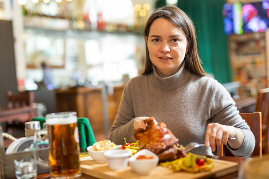 Positive brunette having lunch in Viennese cafe with traditional dish Schweinsstelze, crispy roasted ham hock with vegetable side dishes and glass of beer. Local cuisine