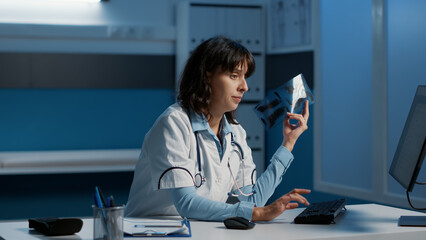 Practitioner holding radiography analyzing lungs before writing medical expertise on computer, planning health care treatment to cure patient disease. Medic working over hours in hospital office