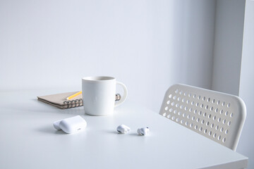 White cup with coffee, notepad, pencil and wireless headphones on a white table.