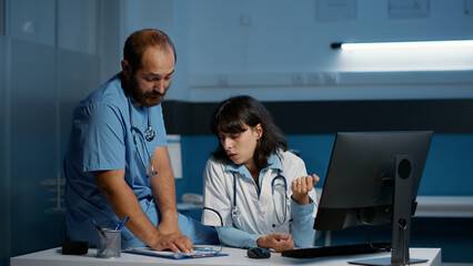 Practitioner doctor standing at desk with assistant analyzing medical expertise while discussing patient disease report. Clinical team working night shift in hospital office. Medicine service