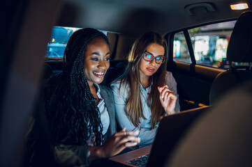 Two multiracial business woman riding in a car and using a laptop