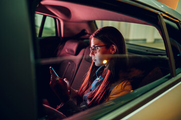 Business woman using smartphone while sitting in a backseat of a car at night