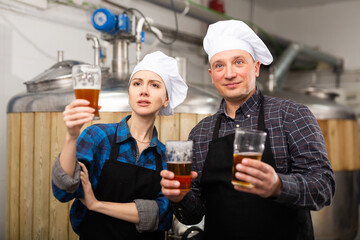 Portrait of man and woman brewmasters in aprons standing in brewery and holding beer glasses.