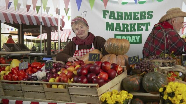 Woman farmer stands at the stall with fresh colorful fruits and vegetables, looks at camera. Farmers market festival. Vegetarian, organic and healthy food. Agriculture.