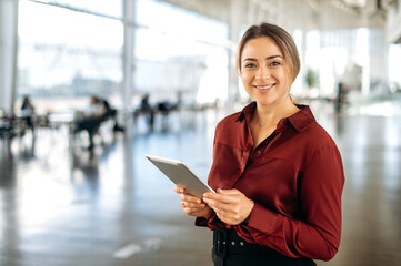 Portrait of positive beautiful intelligent caucasian business lady, recruiter, broker, company employee, in elegant clothes, holding a tablet in her hands, stand in the office, looks at camera, smiles