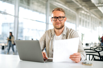 Friendly motivated successful caucasian man, with glasses, IT specialist, company seo, manager, freelancer, sits at a desk with a laptop in coworking, works with documents, looks at camera, smiles