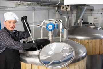 Skilled owner of small craft brewery mixing raw materials in fermentation vessel in workshop..