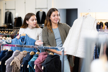 Obraz premium Two cheerful female friends comparing warm white and blue gilets in a shopping mall