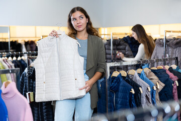 Young positive woman shopping for warm casual clothing in modern store, choosing down vest