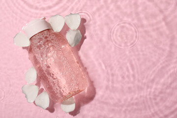 Wet bottle of micellar water and petals on pink background, top view. Space for text