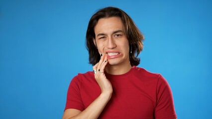 Portrait of Latino Hispanic gender fluid young hipster man 20s with tooth ache problem in mouth isolated on blue background in studio