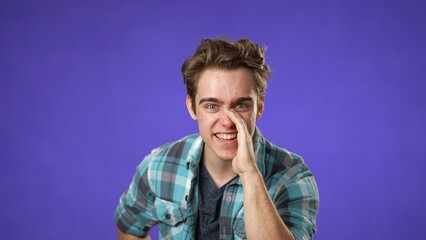 Portrait of caucasian hipster man 20s looks around put hand to mouth tells secret isolated on purple background studio portrait