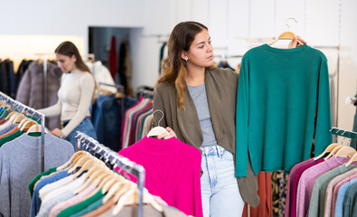 Thoughtful young female choosing between raspberry and green sweaters in a clothing store