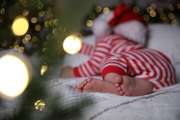 Baby in Christmas pajamas and Santa hat sleeping on bed indoors, focus on feet