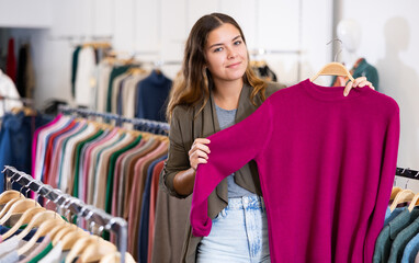 European young female is thinking about buying a raspberry sweater while walking through a clothing store