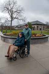 Married senior gay couple are out for a walk in a park on a cold autumn day with cloudy skies.  One man is in a wheelchair with a blanket on his lap whilst his husband pushes him in the wheelchair.