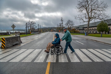 An elderly gay man pushes his husband in a wheelchair in a pedestrian crossing across the road. ...