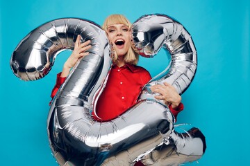 a happy, enthusiastic woman in a red shirt stands on a blue background and holds inflatable balloons in the shape of the number twenty-two in silver color. Horizontal Studio Photography