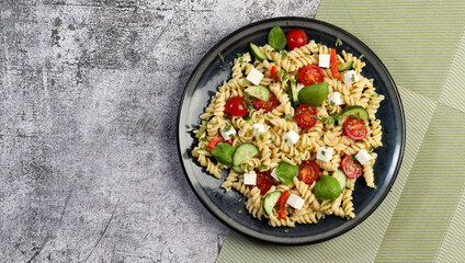 Pasta Salad with tomatoes, bell peppers and feta cheese on a round plate on a dark gray background. Top view, flat lay