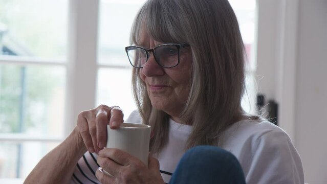 Close up of senior Caucasian woman drinking cup of coffee while working. Backlit shot of older retired woman holding mug of tea. Slow motion 4k