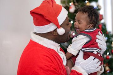 African father wears Santa hat playing with his cute baby on Christmas tree background, lifestyle...