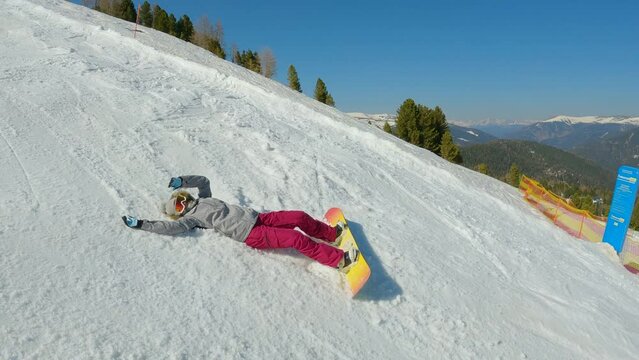 Girl snowboarder falls back on snow while learning to snowboard on ski slope. Young woman landing in snow after losing balance while snowboarding down the piste at snow-covered mountain ski resort.
