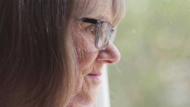 Close up side view of senior woman with glasses looking out window. Moody dramatic close-up of retired older Caucasian lady. Slow motion 4k